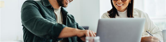 Een man en een vrouw zitten samen samen met plezier achter een laptop. De man wijst wat aan op het scherm, de vrouw maakt een aantekening.