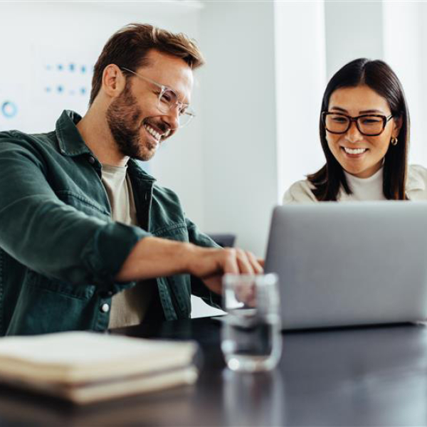 Een man en een vrouw zitten samen samen met plezier achter een laptop. De man wijst wat aan op het scherm, de vrouw maakt een aantekening.