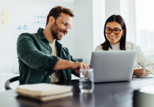 Een man en een vrouw zitten samen samen met plezier achter een laptop. De man wijst wat aan op het scherm, de vrouw maakt een aantekening.
