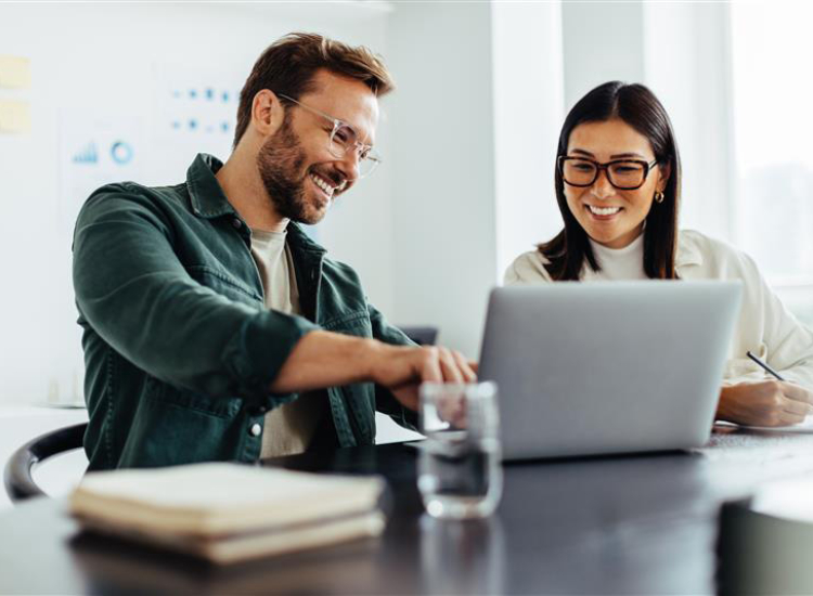 Een man en een vrouw zitten samen samen met plezier achter een laptop. De man wijst wat aan op het scherm, de vrouw maakt een aantekening.