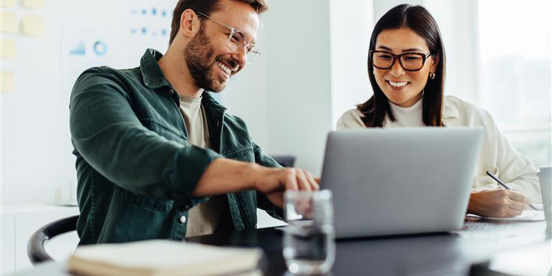 Een man en een vrouw zitten samen samen met plezier achter een laptop. De man wijst wat aan op het scherm, de vrouw maakt een aantekening.